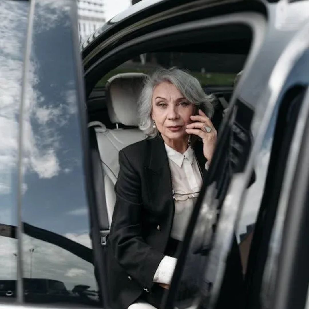 A confident woman in a black blazer talking on the phone by a car.
